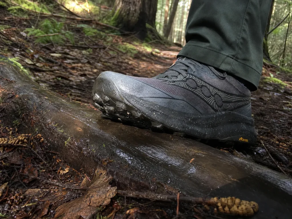 Sticky rubber outsole gripping a wet root crossing the trail, hiking shoe edge flexed over the curve