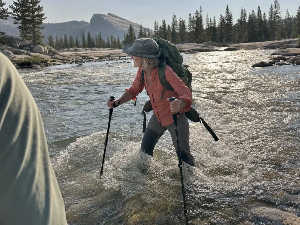 Hiker facing upstream with poles planted and hip belt unbuckled while fording a Sierra Nevada snowmelt creek