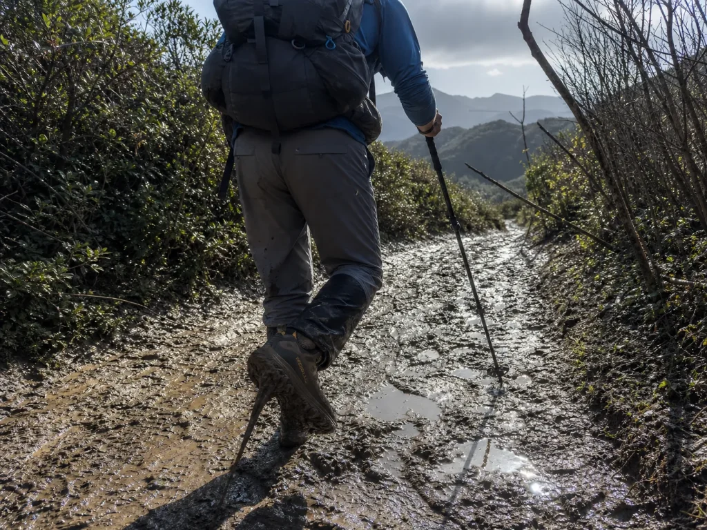 Backpacker in low-cut hiking shoes stepping through mud under a loaded pack on a graded trail