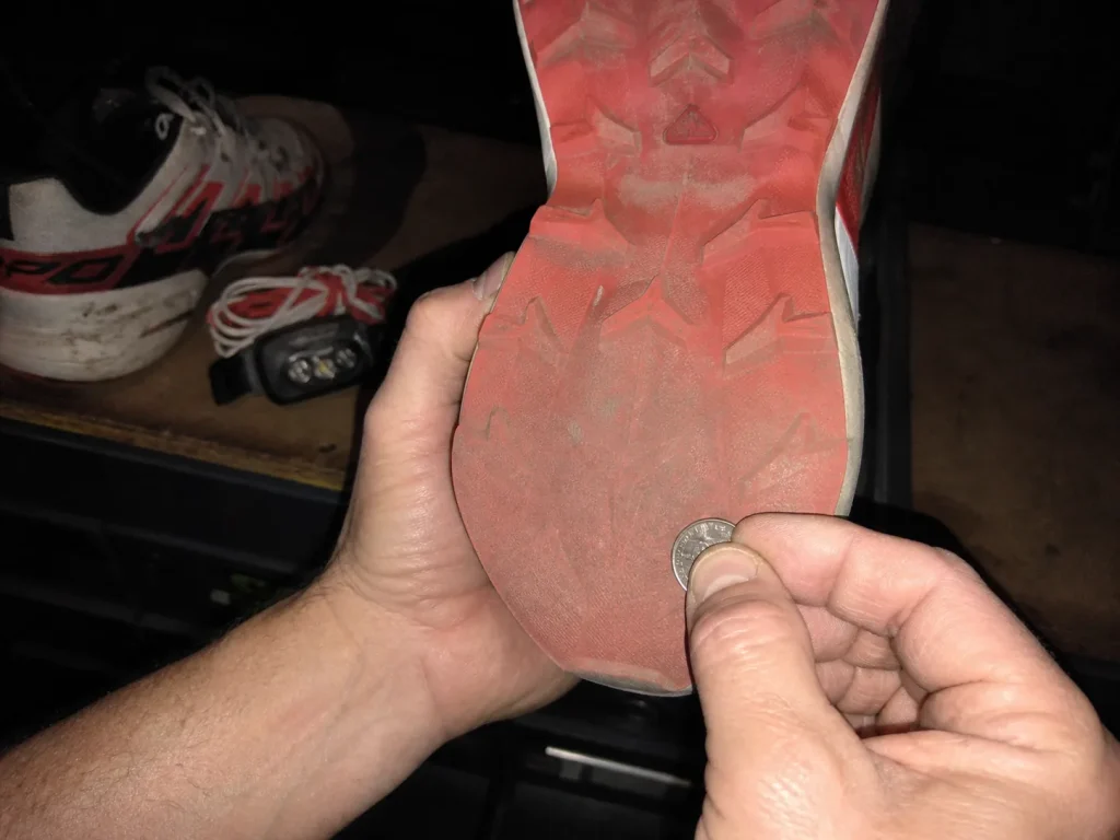 Hiker checking worn heel lug depth with a coin edge in a garage the night before a muddy hike