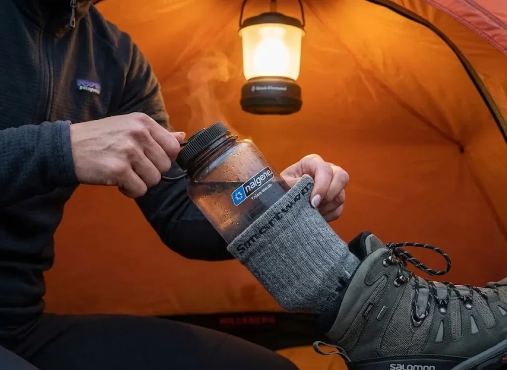 A hiker inside a tent placing a hot water bottle inside a Smartwool sock to dry a Salomon hiking boot safely.