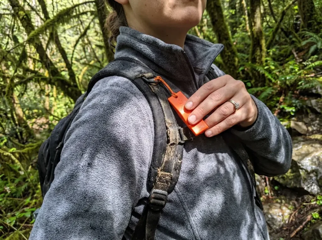 A flat pealess rescue whistle clipped to a backpack shoulder strap for fast emergency access on the trail