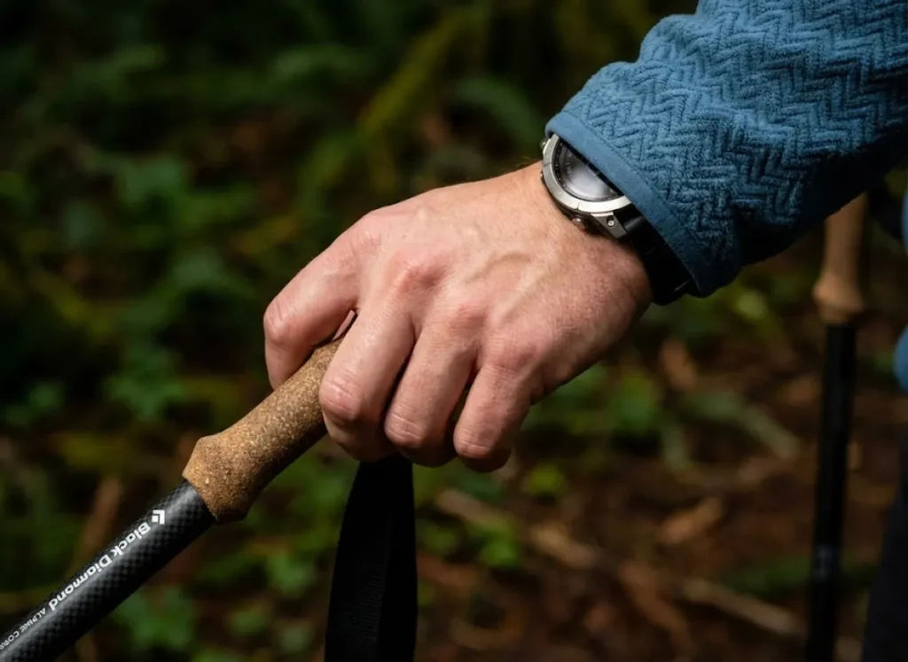 Close-up of a hiker's hand holding a Black Diamond trekking pole and wearing a Garmin watch, analyzing the trail surface with precision.