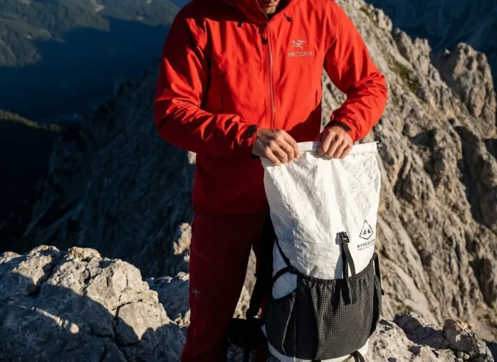 A professional hiker on a rocky ridge wearing an Arc'teryx jacket and Hyperlite Mountain Gear backpack, demonstrating perfect posture and balance against a dramatic alpine sky.