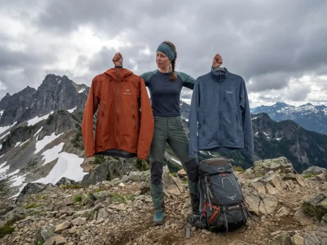 hardshell-vs-softshell-hiking-jacket-comparison Hiker comparing hardshell and softshell jackets at alpine ridge pass with mountain views