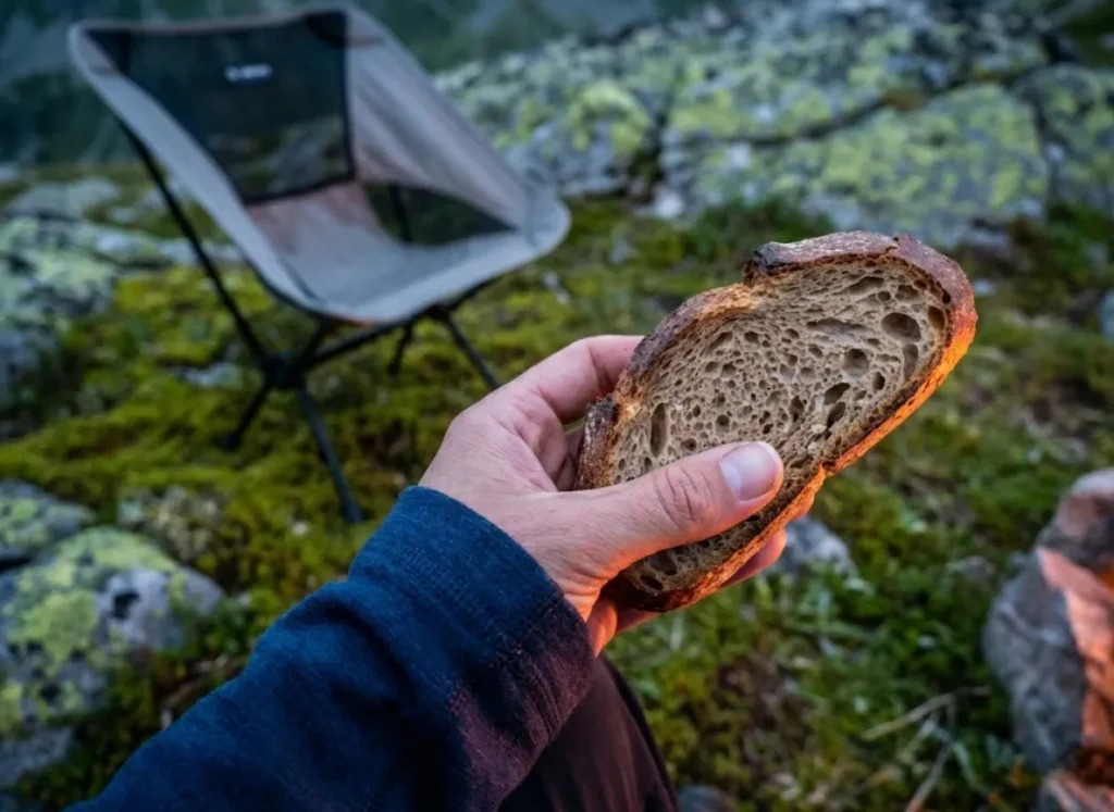 Close-up POV shot of a perfectly baked slice of camp bread with a crisp crust, held by a hiker wearing Smartwool gear in a campsite setting.