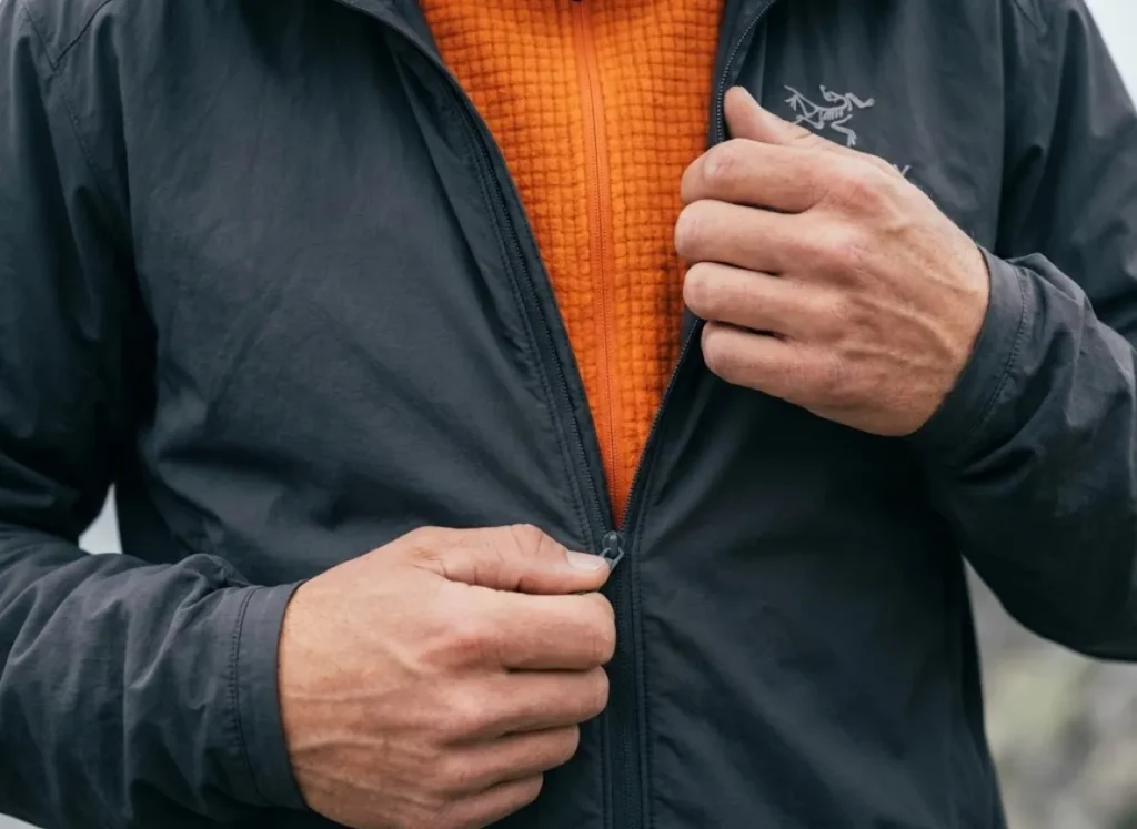 Close-up of a hiker unzipping a wind shell to reveal a bright orange grid fleece mid-layer, demonstrating mechanical venting.