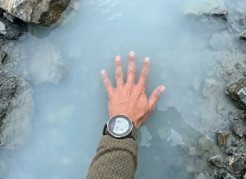 POV shot of a hiker's clean hand performing a turbidity knuckle test in a glacial stream, wearing a Suunto watch and Patagonia fleece.