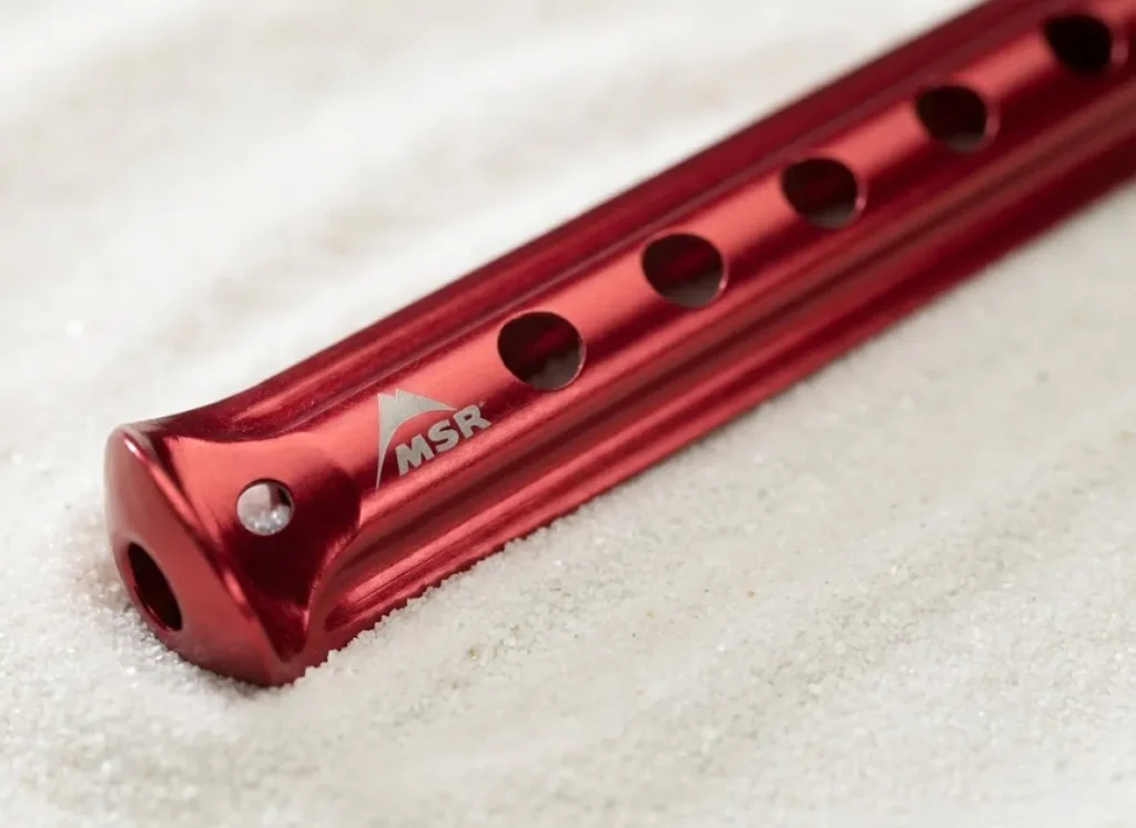 Macro photography of a red anodized MSR Blizzard tent stake lying on white sand, highlighting its concave surface area and aluminum texture.