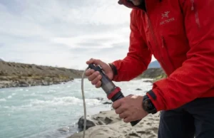 Pre Filter Silty Water to Save Your Pump Filter Alpine guide in Arc'teryx gear kneeling by a glacial river, preparing a pristine MSR Guardian water pump filter against a backdrop of turquoise silty water.