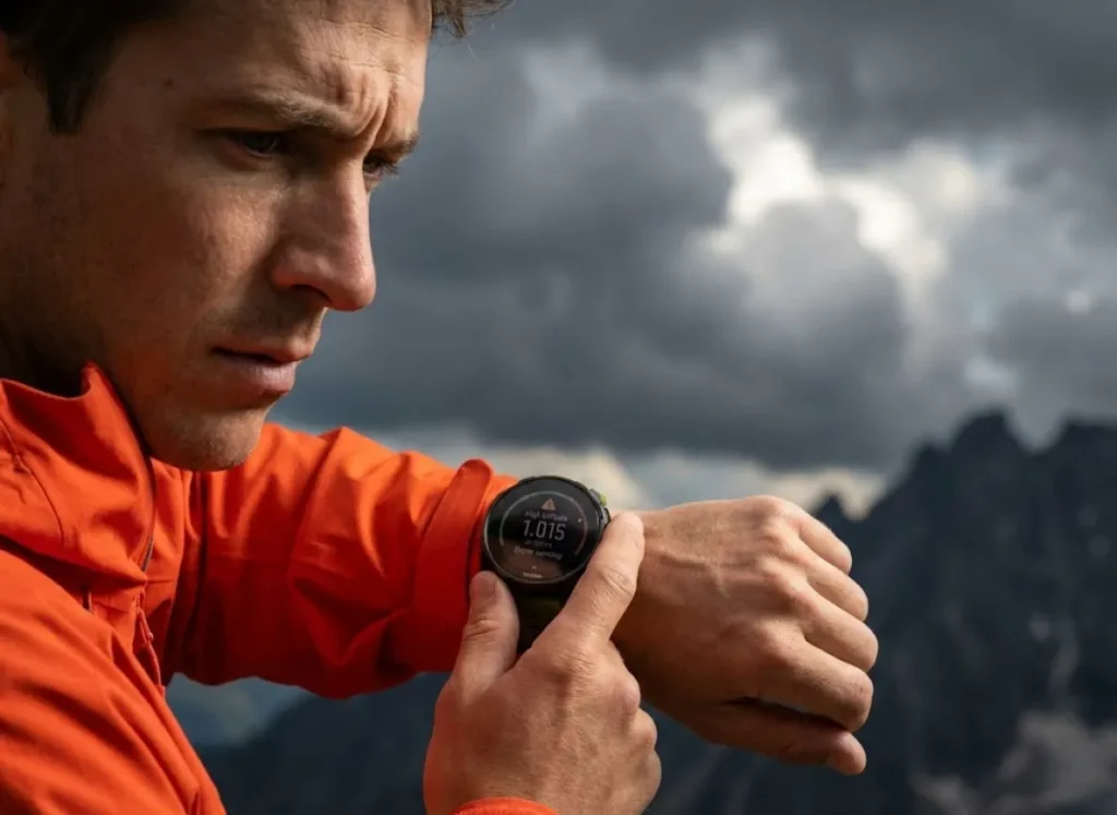 A mountaineer checking a Suunto watch for altitude and weather alerts, with storm clouds building in the background, depicting the decision to turn around.