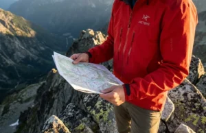 Read Terrain Features to Orient Without a Compass A professional hiker in an Arc'teryx jacket standing on a high mountain ridge, consulting a topographic map against a backdrop of dramatic peaks during golden hour.