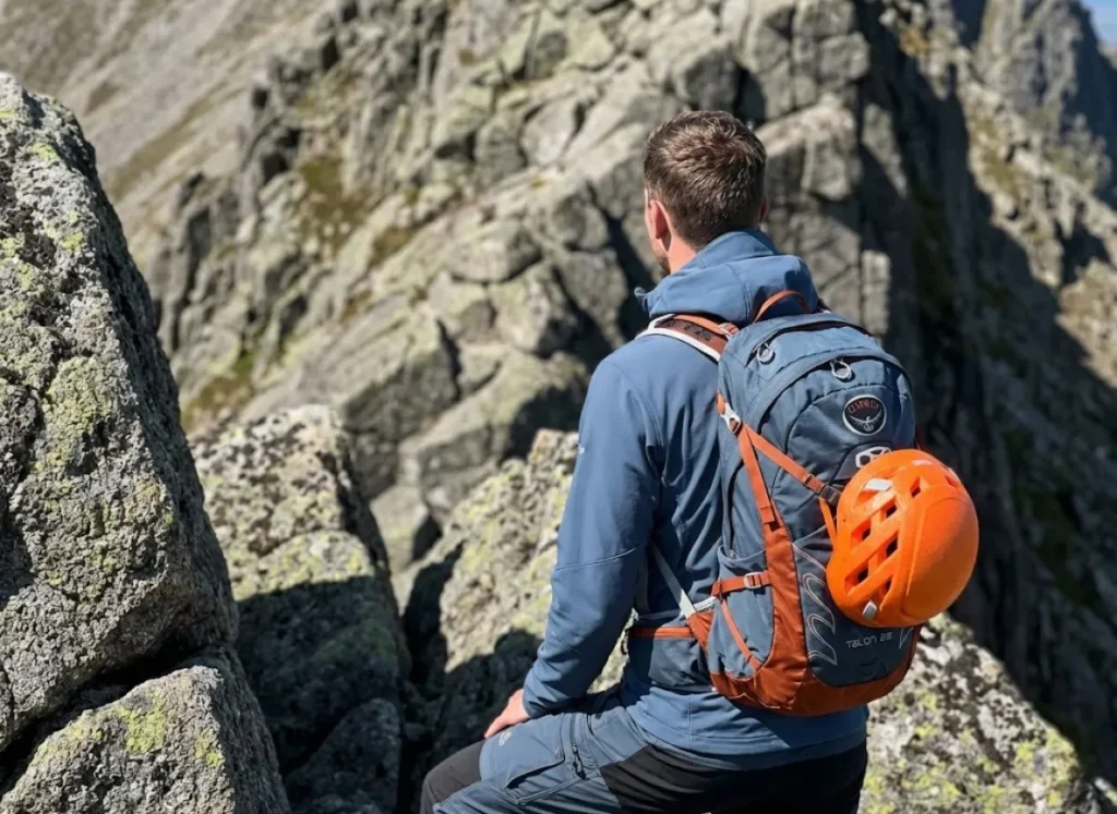 A climber with an Osprey backpack looks up at a steep, trail-less Class 3 ridge, visualizing the route ahead.