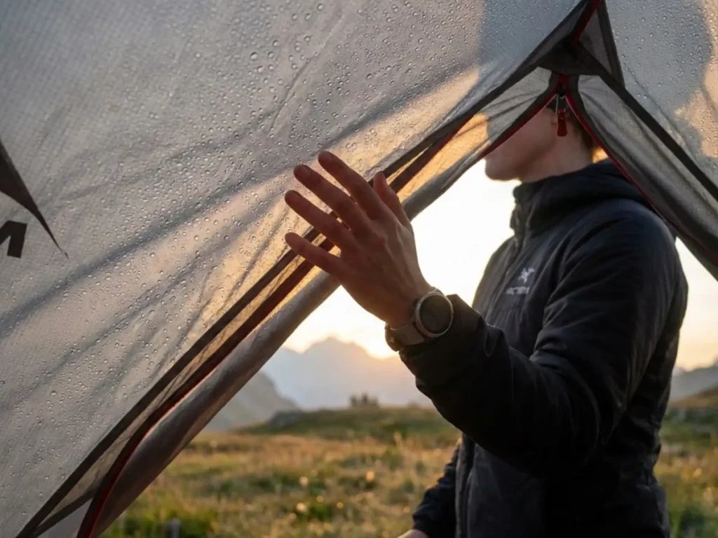 Backpacker examining tent rainfly condensation droplets at alpine sunrise campsite