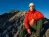 Transition From Hiking to Peak Bagging Safely A professional mountaineer wearing an Arc'teryx jacket and Black Diamond helmet stands at the edge of a trail looking up at a rocky peak, illuminated by golden hour sunlight.