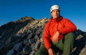 Transition From Hiking to Peak Bagging Safely A professional mountaineer wearing an Arc'teryx jacket and Black Diamond helmet stands at the edge of a trail looking up at a rocky peak, illuminated by golden hour sunlight.
