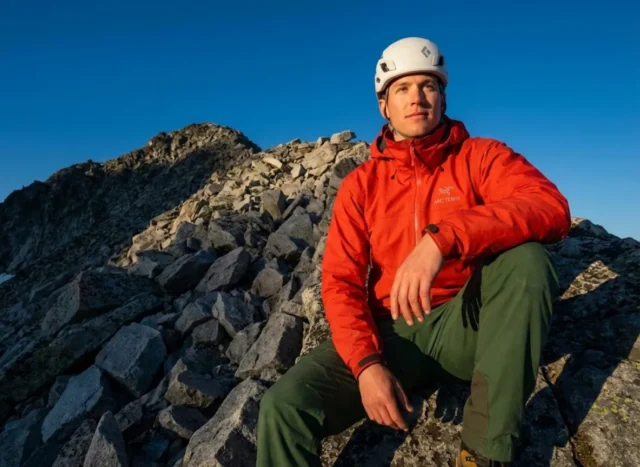 transition-hiking-peak-bagging-safely A professional mountaineer wearing an Arc'teryx jacket and Black Diamond helmet stands at the edge of a trail looking up at a rocky peak, illuminated by golden hour sunlight.