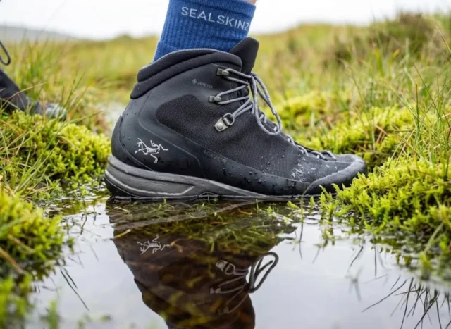 waterproof-socks-hiking-bog-main Close-up of a high-end hiking boot stepping into water, highlighting the texture of a waterproof sock cuff and water beading technology.