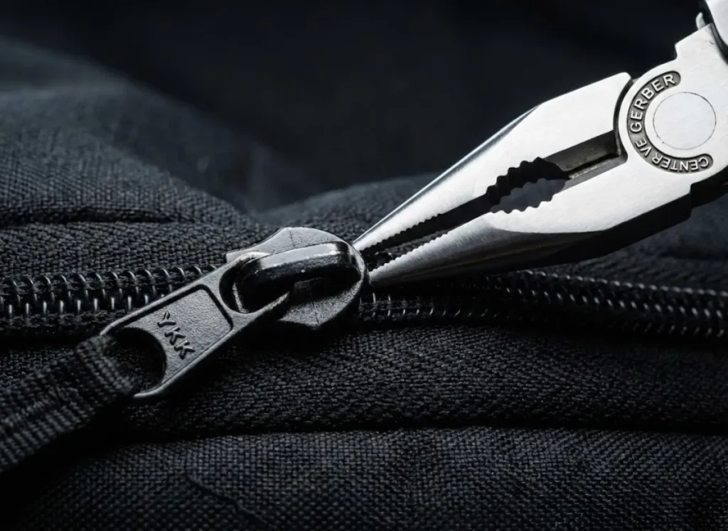 Macro photography view of needle-nose pliers gently crimping the back of a YKK zipper slider to fix a separation.