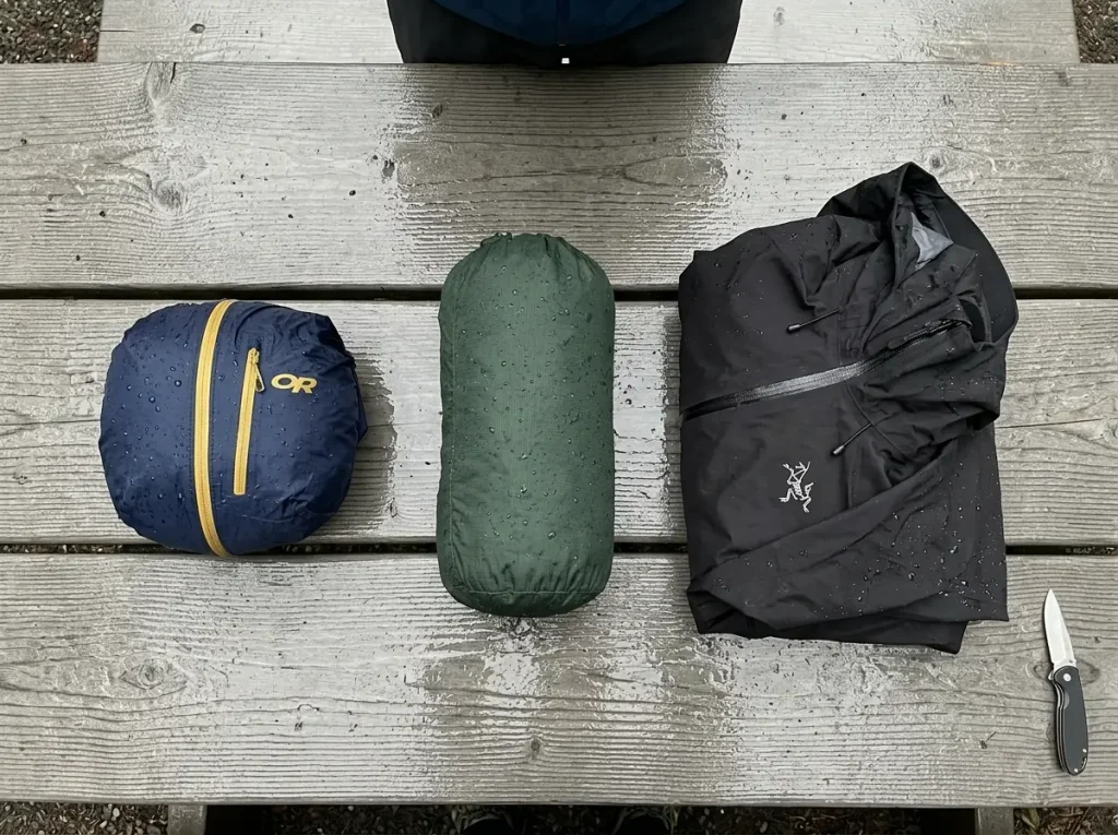 Three rain jackets stuffed to different sizes lined up on a wet picnic table from ultralight to three-layer