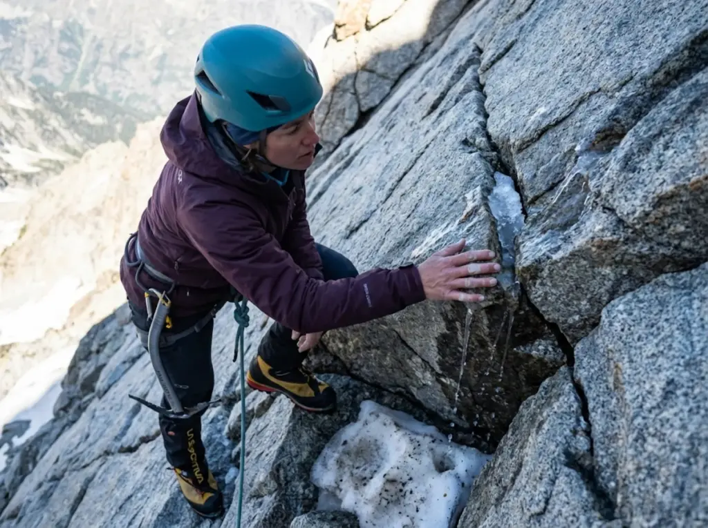 Alpinist examining ice-filled rock fracture in alpine terrain showing freeze-thaw destabilization