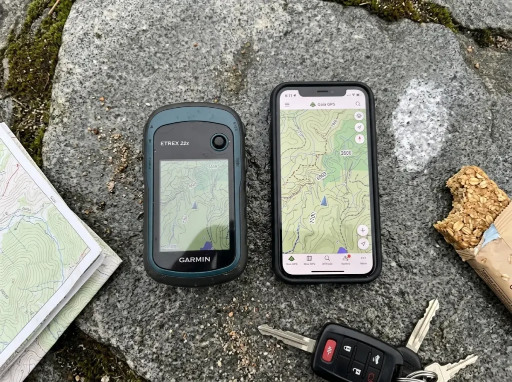 A handheld GPS unit and a smartphone laid side by side on a rock comparing hiking navigation options