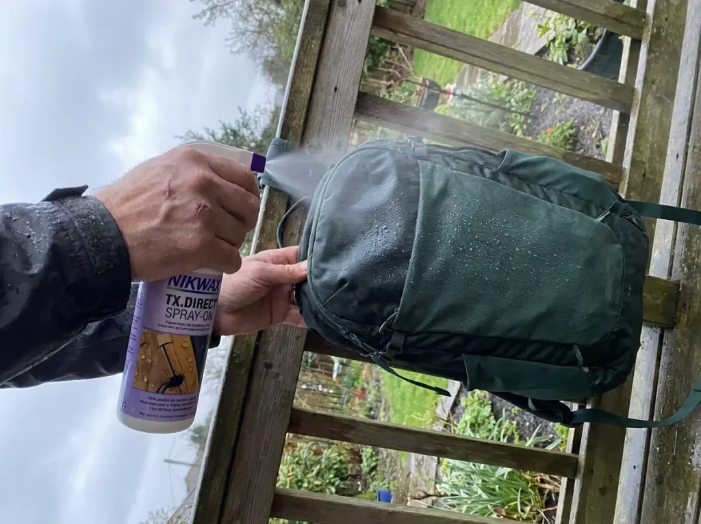 Hands spraying Nikwax DWR water repellent onto a backpack panel to refresh its waterproofing