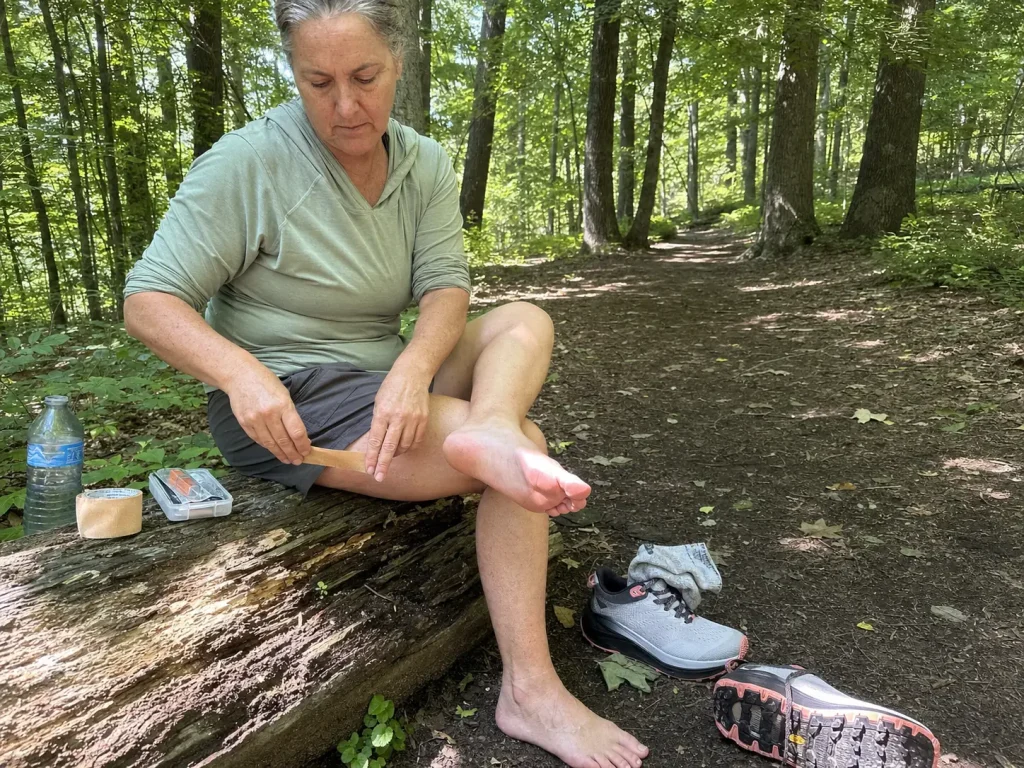 Hiker taping a hot spot on her foot at a trail break beside her hiking shoes