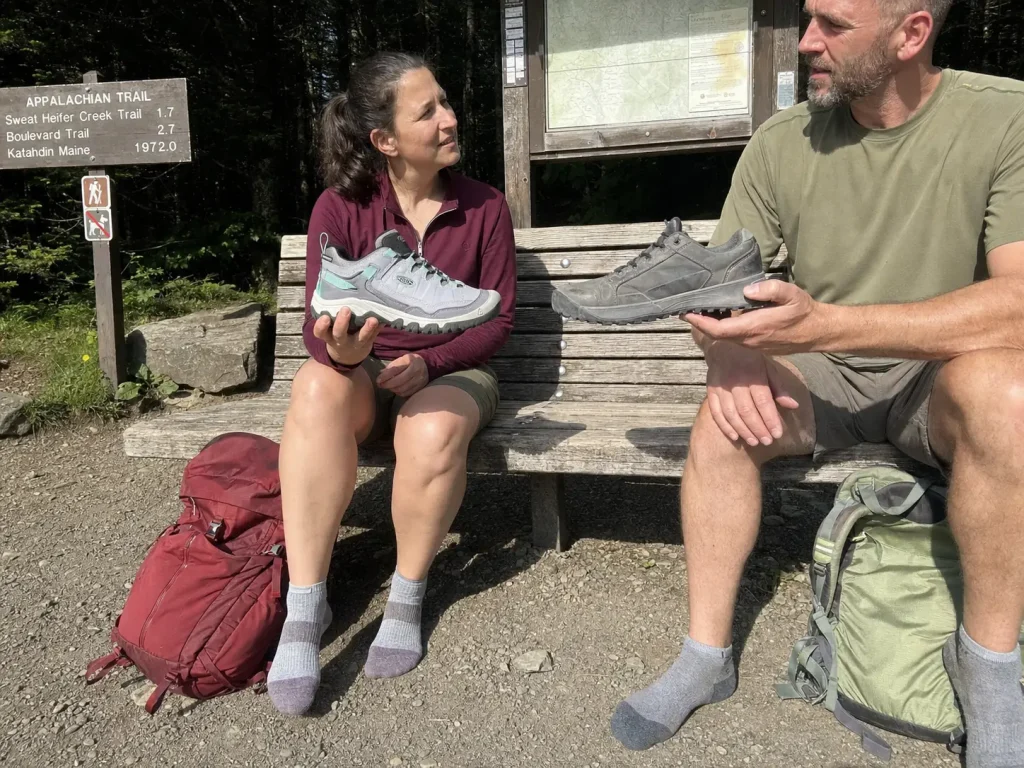 Two hikers comparing men's and women's wide-fit hiking shoes at a trailhead bench