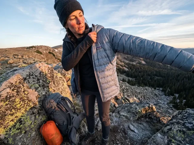 best-lightweight-down-jacket-for-hiking-hero Hiker pausing at ridgeline to pull on lightweight down jacket in cold alpine morning light