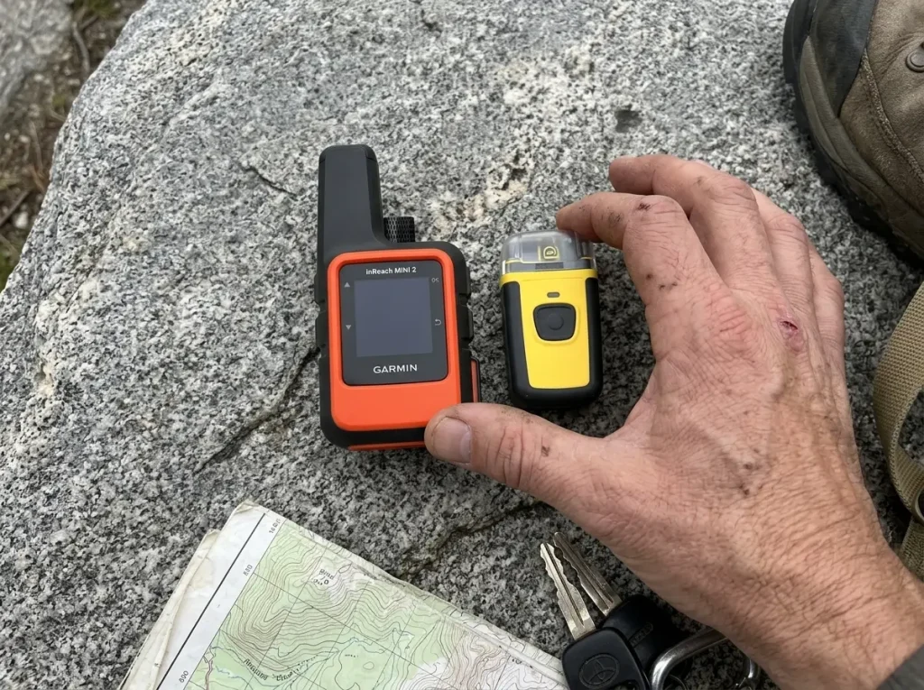 Hiker comparing a Garmin inReach Mini 2 messenger and a compact PLB laid on a granite rock