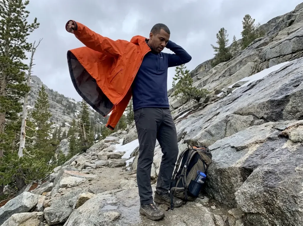 Hiker putting on a separate rain shell over a fleece on a mountain trail