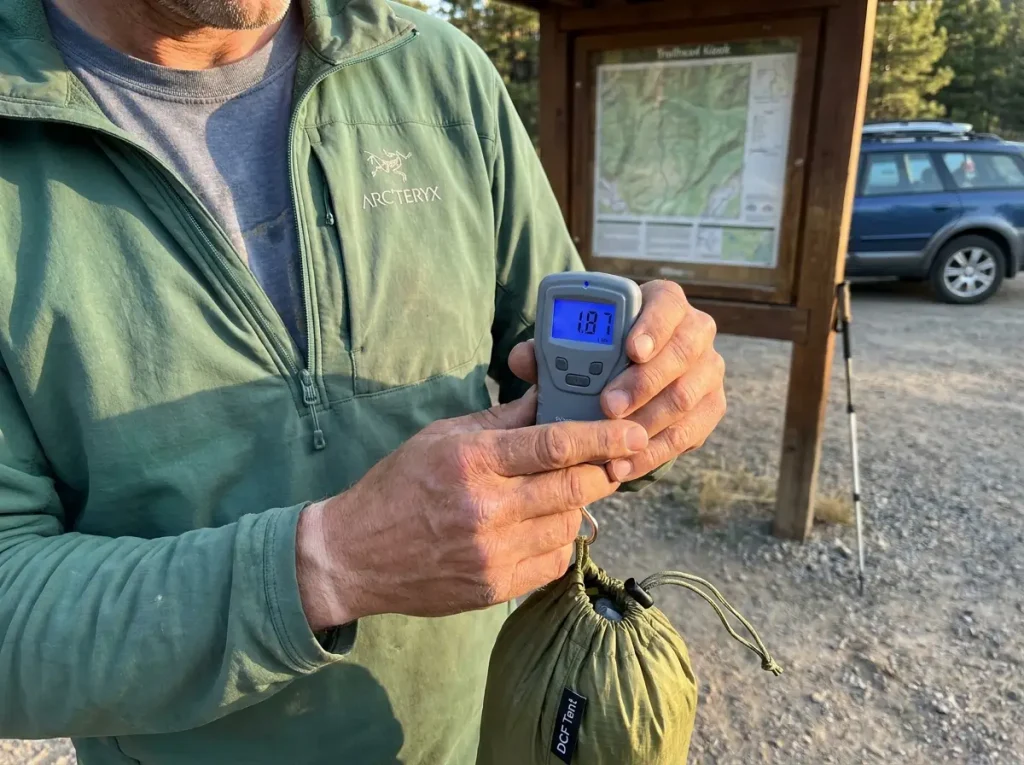 Hiker weighing a packed ultralight tent on a hanging scale at a trailhead