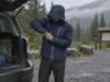 Rain Jacket Sizing for Layering Without Guessing Hiker pulling a rain jacket over a fleece at a wet trailhead, testing rain jacket sizing for layering