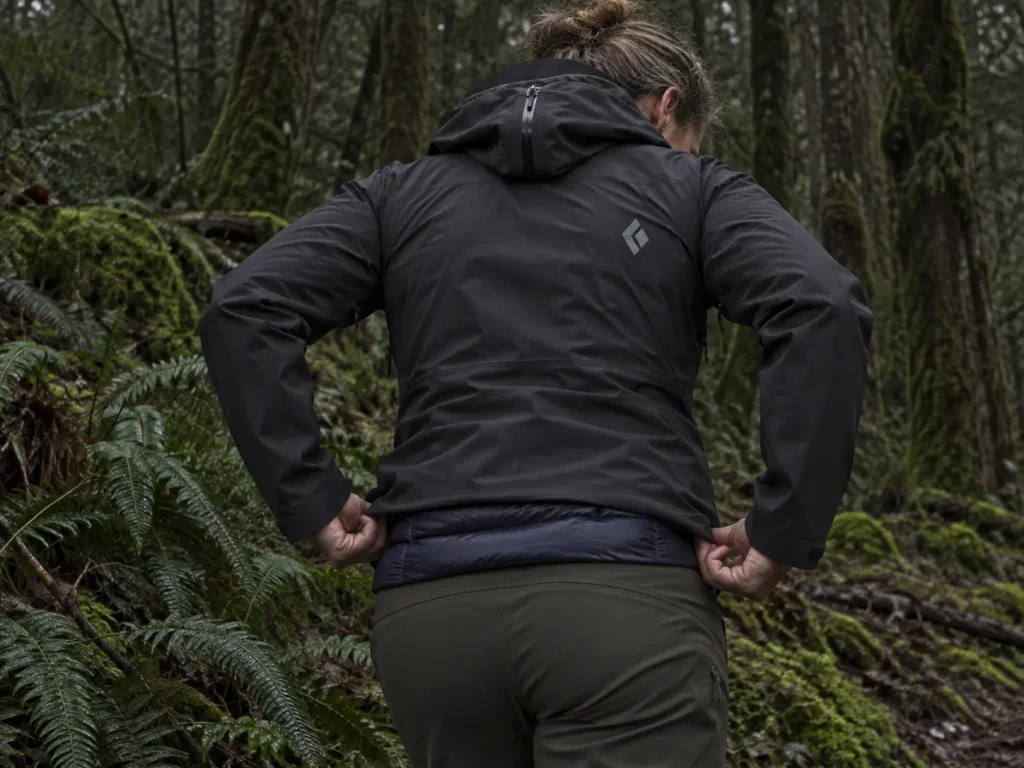 Hiker's rain shell pulling tight across the shoulders over a puffy jacket on a cold trail stop