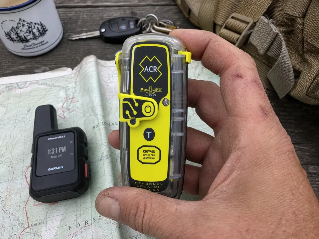 Hand holding an ACR ResQLink personal locator beacon next to a satellite messenger on a map