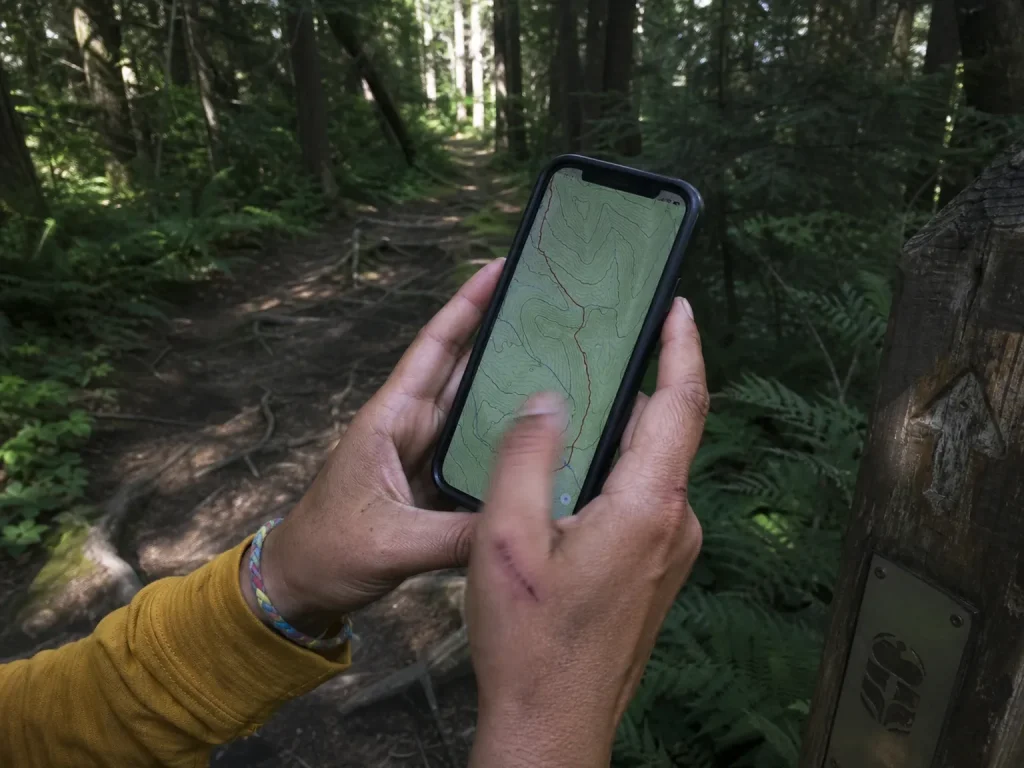 Close-up of a hiker's hands holding a phone showing a trail map beside a wooden trail marker post