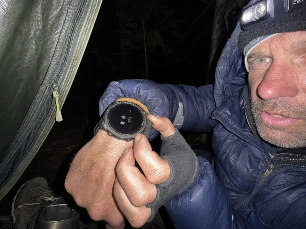 Backpacker checking GPS watch battery by headlamp at dusk camp on a multi-day trip