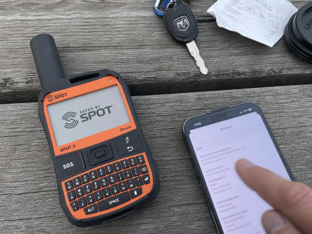 SPOT X satellite messenger beside a phone showing a subscription plan screen on a trailhead bench