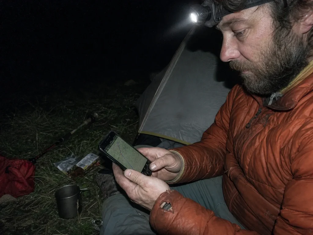 Hiker planning the next day's route on a phone by headlamp at a dusk campsite
