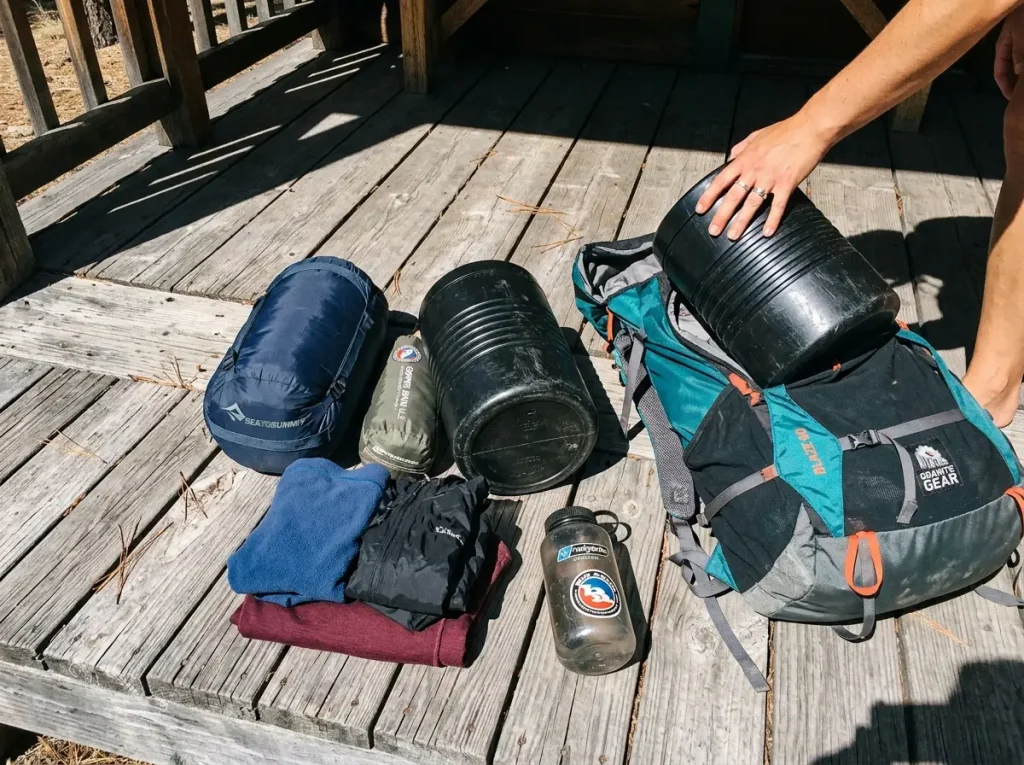 Multi-day backpacking gear laid out beside a 60L pack showing how a bear canister fills volume