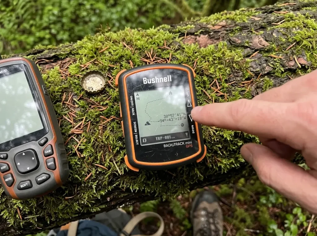 A compact Bushnell handheld GPS and a larger unit set side by side on a mossy log