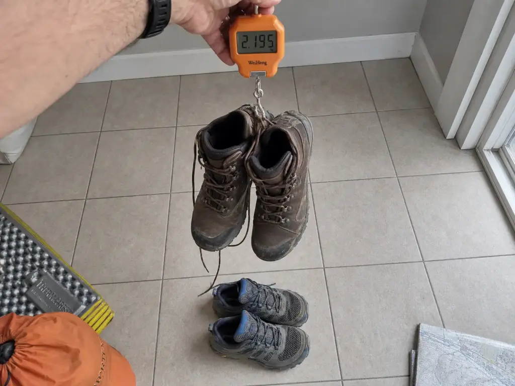 Hiking boots on a luggage scale next to trail runners, weighing backpacking footwear
