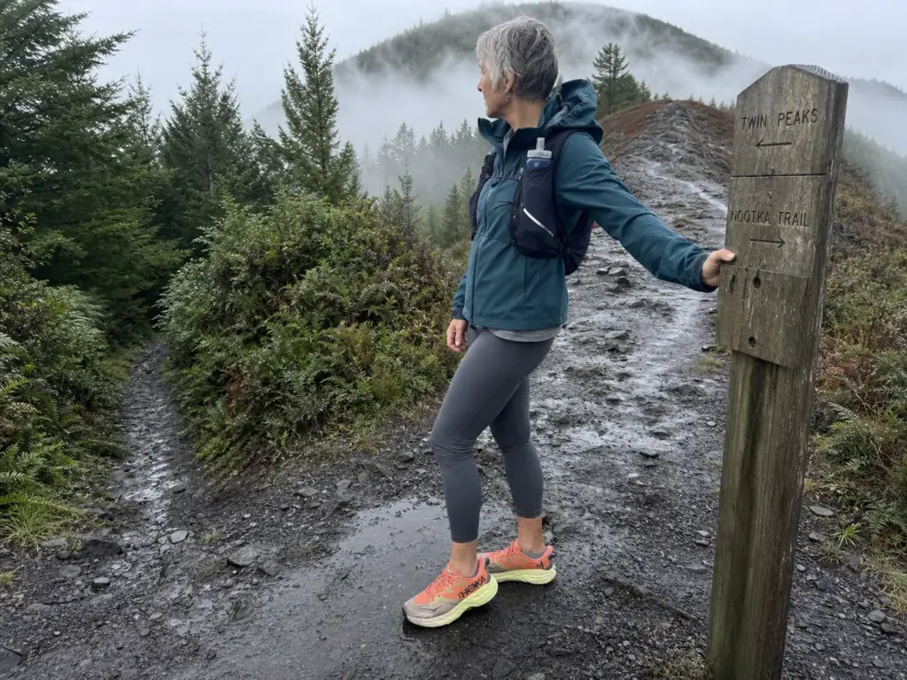 Woman pausing at a forked trail junction sign in lightweight women's hiking shoes on a wet morning