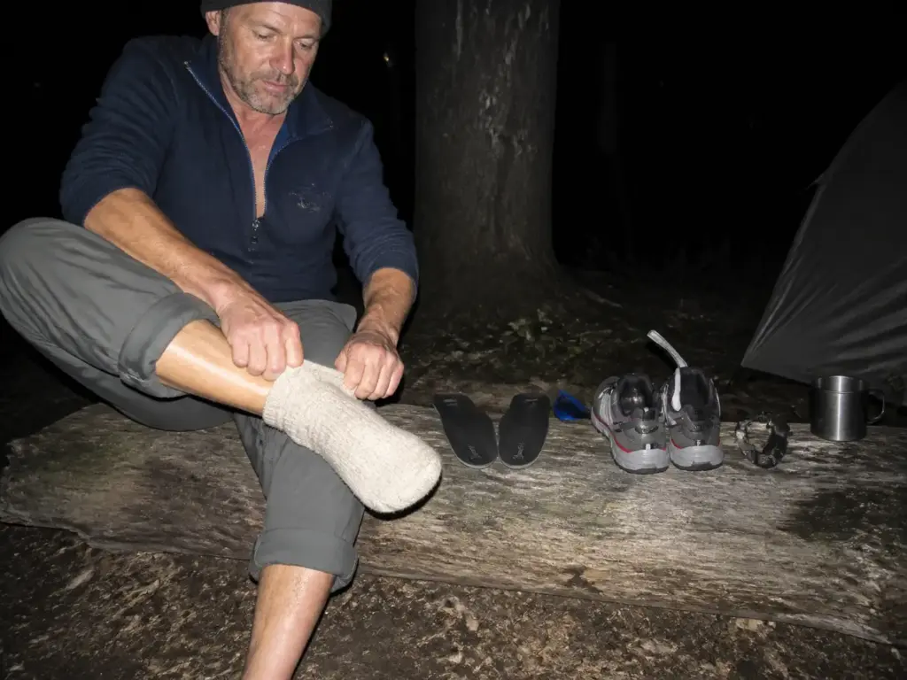 Hiker changing into dry camp socks at dusk beside damp waterproof hiking shoes at a campsite