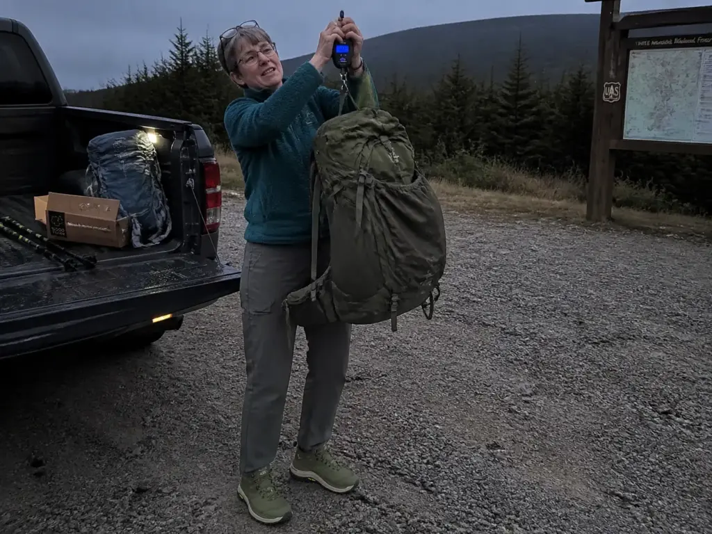 A backpacker weighing a loaded pack at the trailhead before choosing footwear