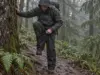 Best Rain Pants for Hiking That Actually Breathe Hiker pulling on rain pants for hiking beside a wet conifer trail under grey rain