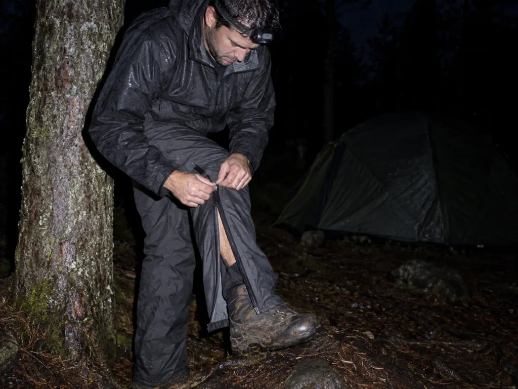 Hiker working a side zip open over muddy boots at dusk beside a tent