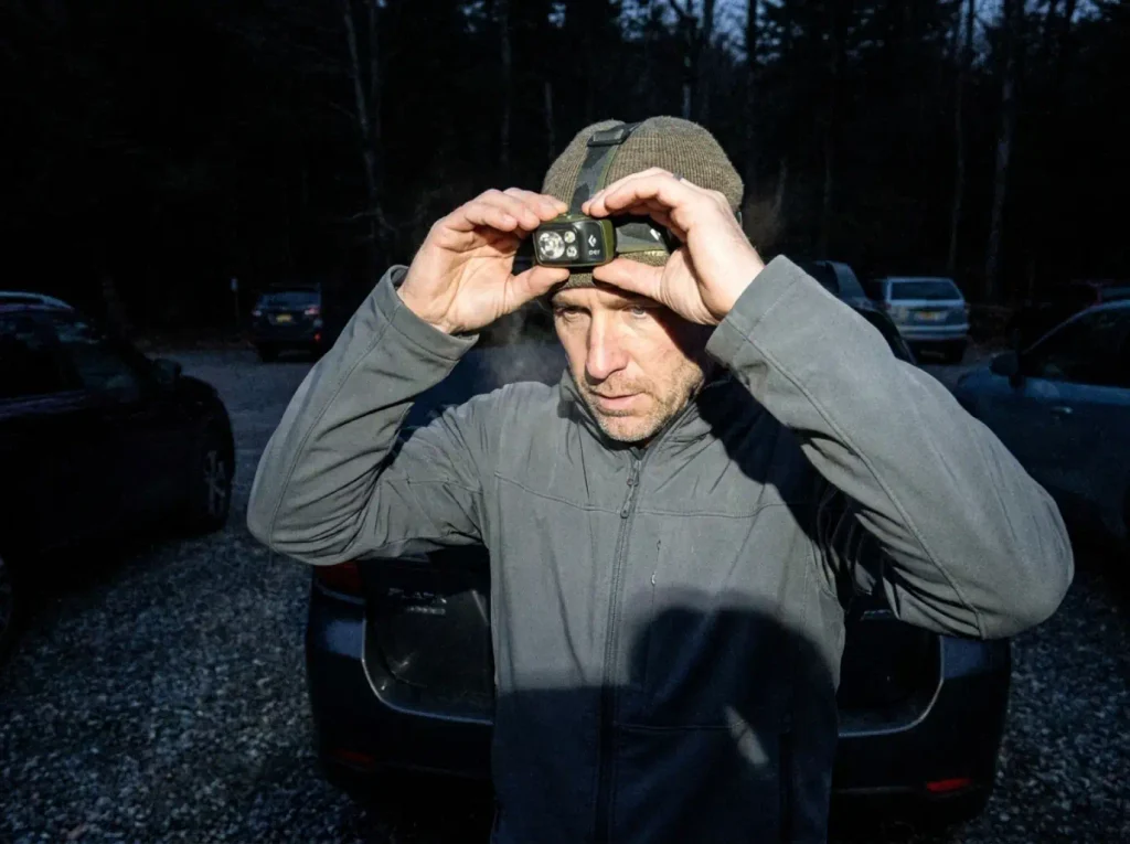 Hiker with a Black Diamond Spot 400 headlamp at a dark Adirondack trailhead lot before dawn