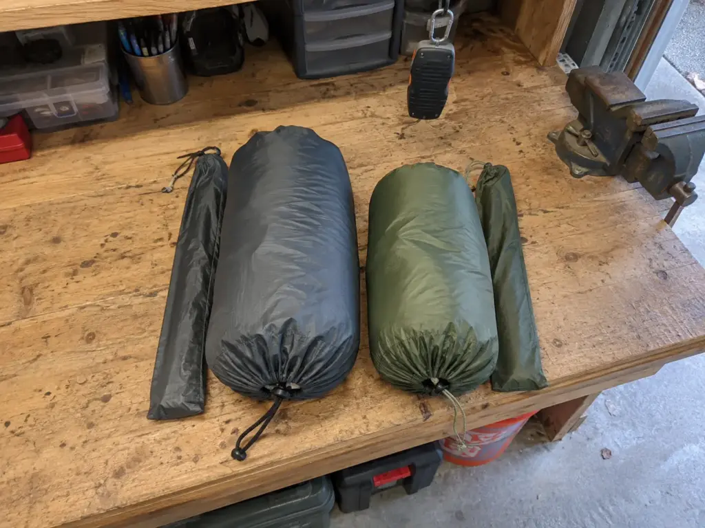 Two packed tent stuff sacks side by side on a garage workbench beside a hanging luggage scale for a tent spec comparison